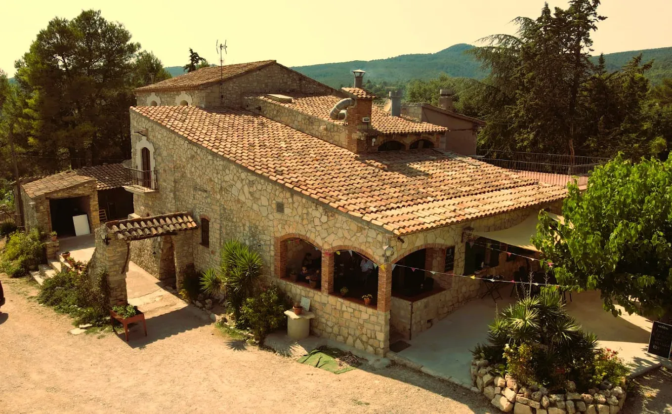 La masia del Mas Pla des de fora: façana de pedra, teulada de teula àrab, porxo d’arcs i la muntanya al darrere